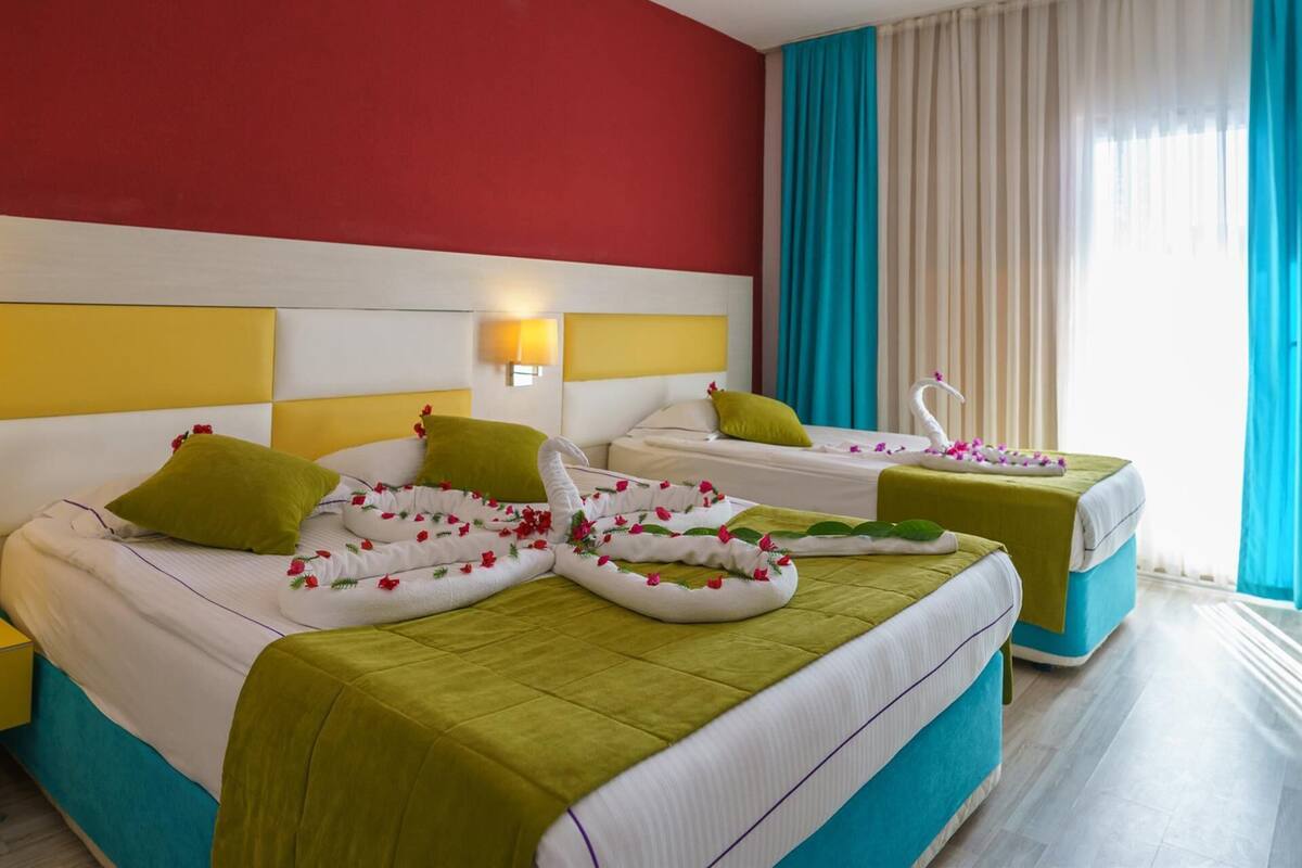 Habitación de hotel con dos camas, una pared roja, un cabecero amarillo y un ventanal con cortinas.