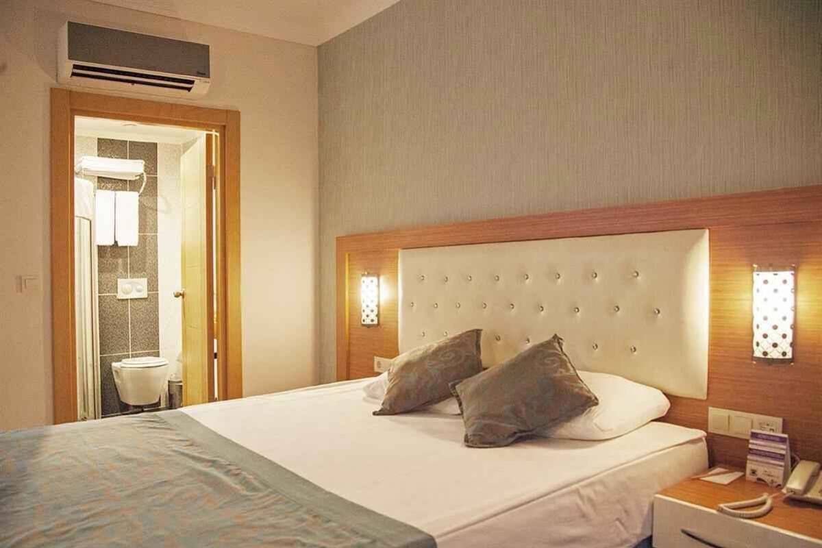 Habitación de hotel con una cama grande, cabecera de madera, dos lámparas de noche, teléfono en la mesita y baño visible a través de una puerta abierta.