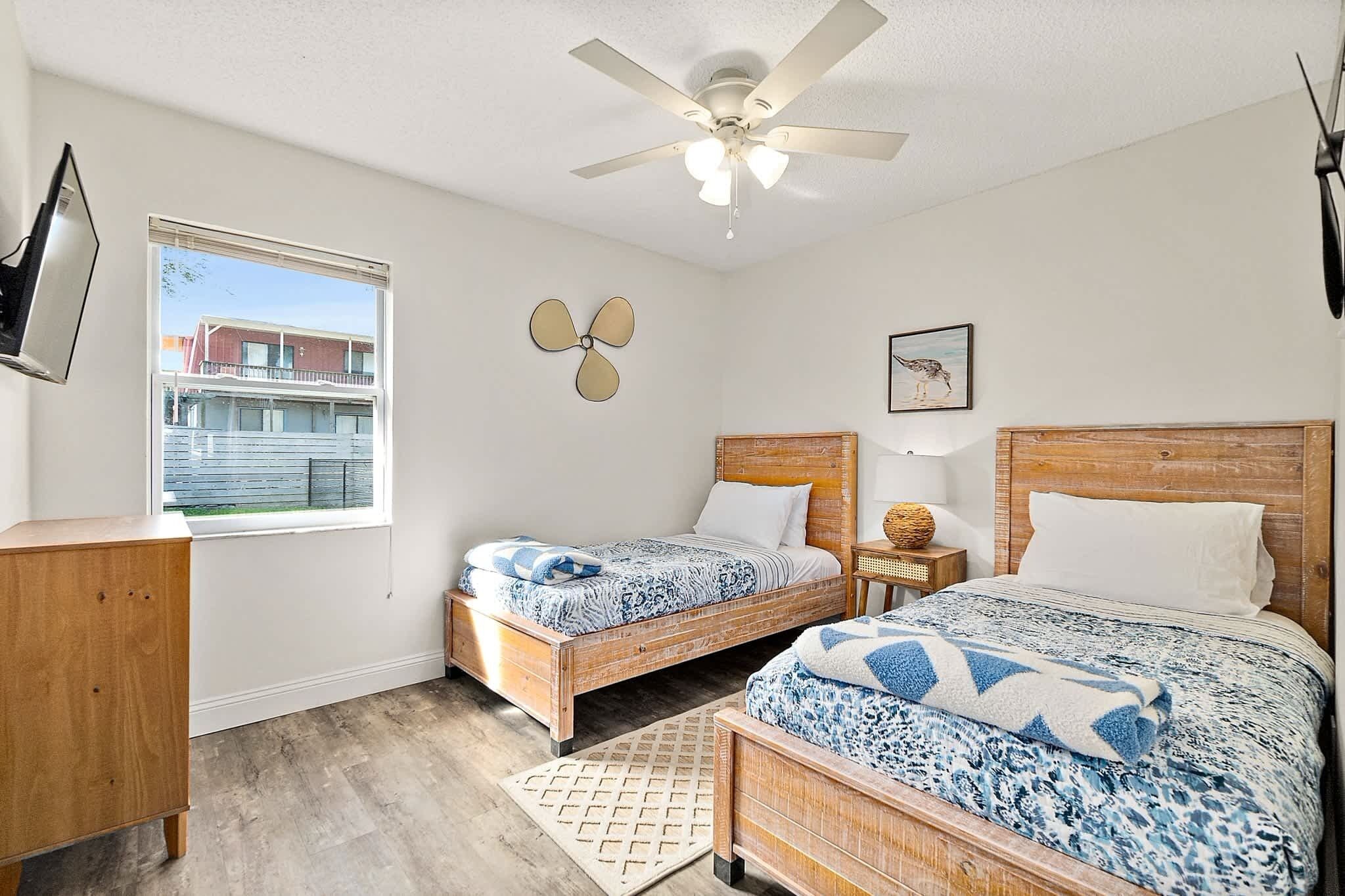 A bedroom with two beds, a ceiling fan, a window with a view, and a wooden dresser.