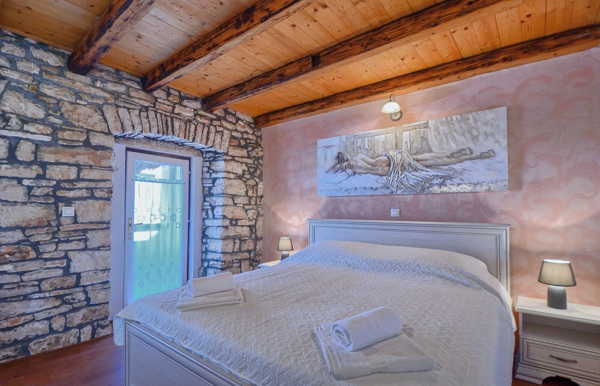 Une chambre avec un mur en pierre, des poutres en bois au plafond, un lit recouvert d’une courtepointe, un tableau accroché au mur et une table de chevet avec une lampe.