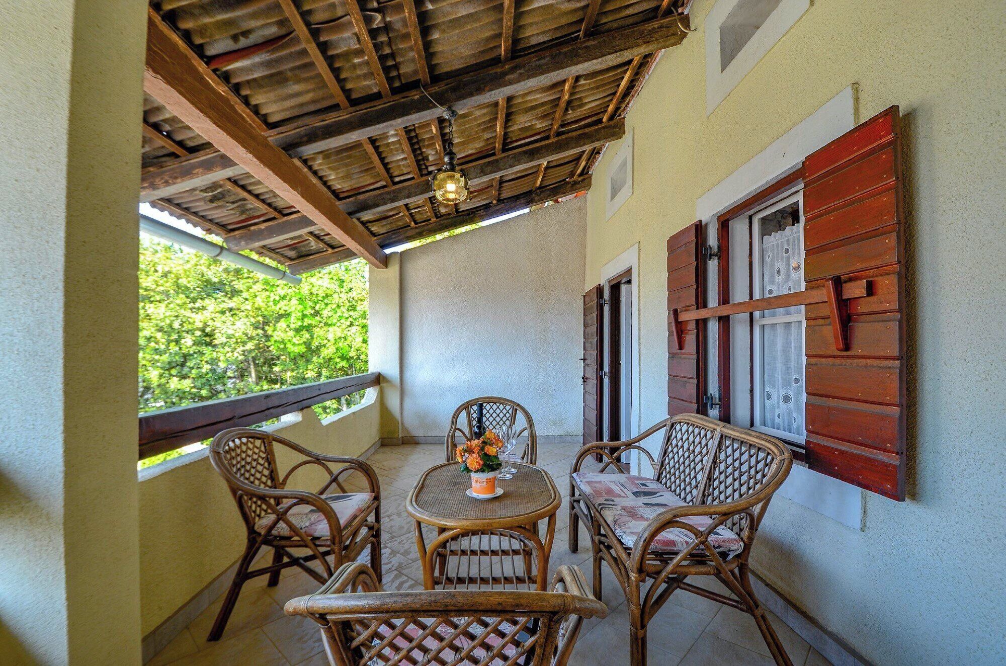 Un espace extérieur aménagé, abrité et doté de meubles en osier et d’une table, une fenêtre avec des volets et une vue sur la verdure environnante.