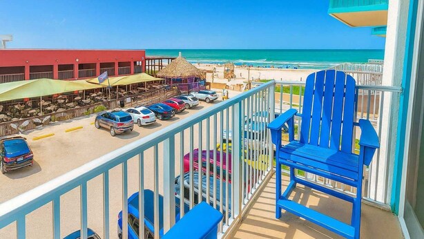 A balcony with a blue chair, a view of the beach, and a row of parked cars.