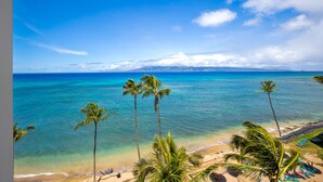 A beach with clear turquoise waters, palm trees, and a view of distant mountains. - Hawaii Life Rentals - Kahana Ocean Front (Lahaina)