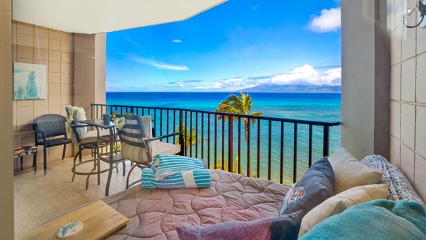 A balcony with a view of the ocean, a dining area with chairs and a table, and a sofa with cushions. - Hawaii Life Rentals - Kahana Ocean Front (Lahaina)