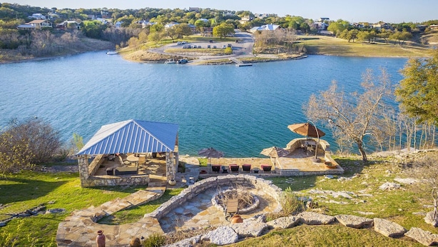 Une construction en pierre surmontée d’un toit métallique, qui surplombe un lac et est entourée de prairies et d’arbres.