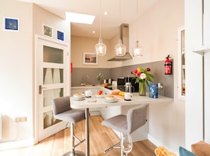A modern kitchen with a dining area, featuring a white countertop, bar stools, a coffee machine, and a fire extinguisher.