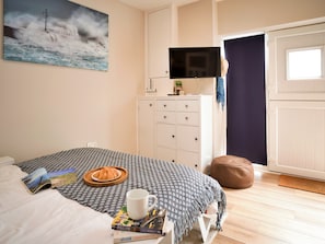 A bedroom with a bed, a bedside table with books and a mug, a dresser, a television, and a view of the outside.