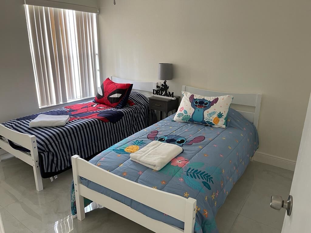 Dos camas individuales con ropa de cama diferente, una mesita de noche con lámpara y una ventana con persianas.