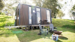 A small, modern mobile home with a deck, outdoor seating, and a fire pit.