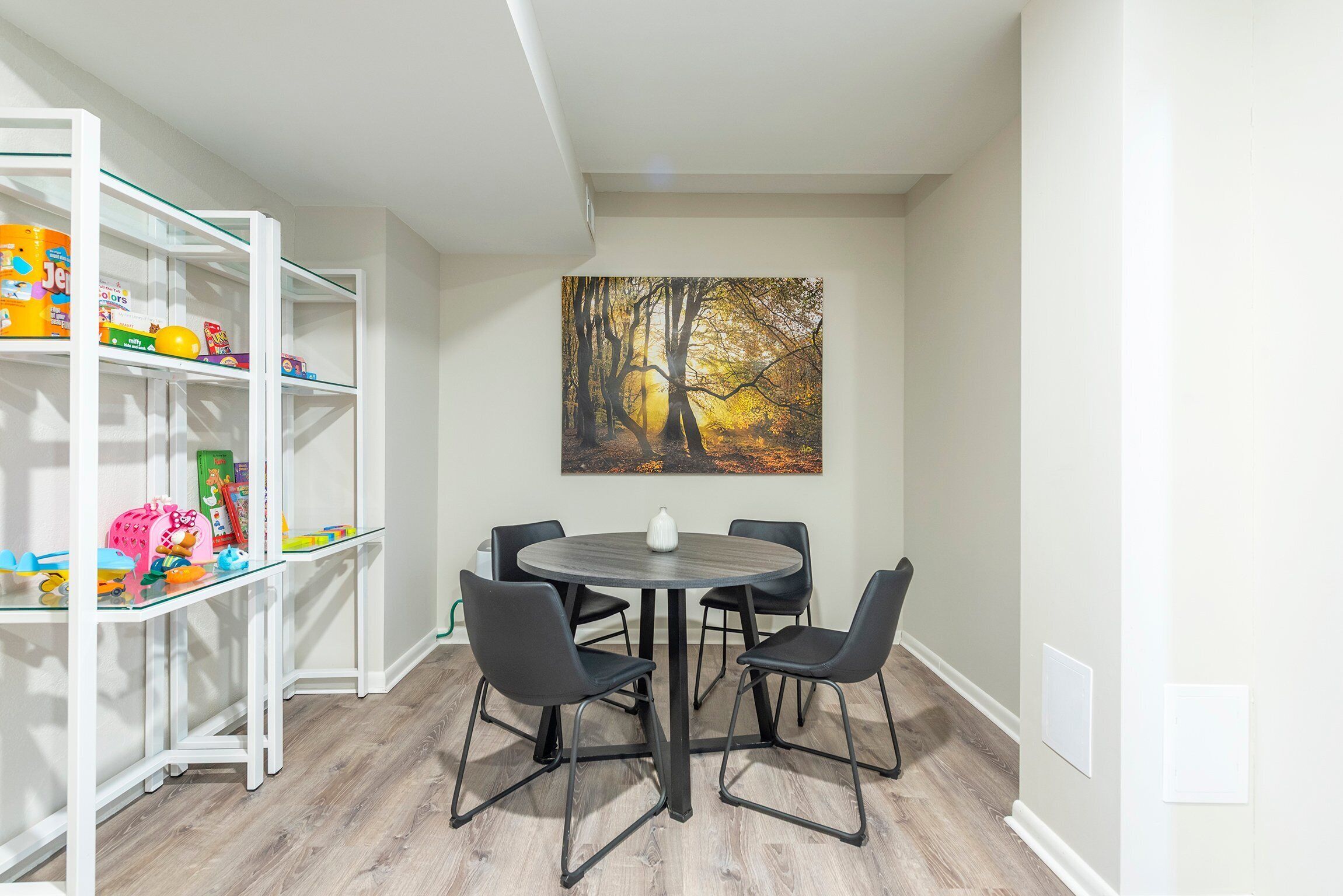 A room with a round table and four chairs, a bookshelf with toys, and a painting of a forest.
