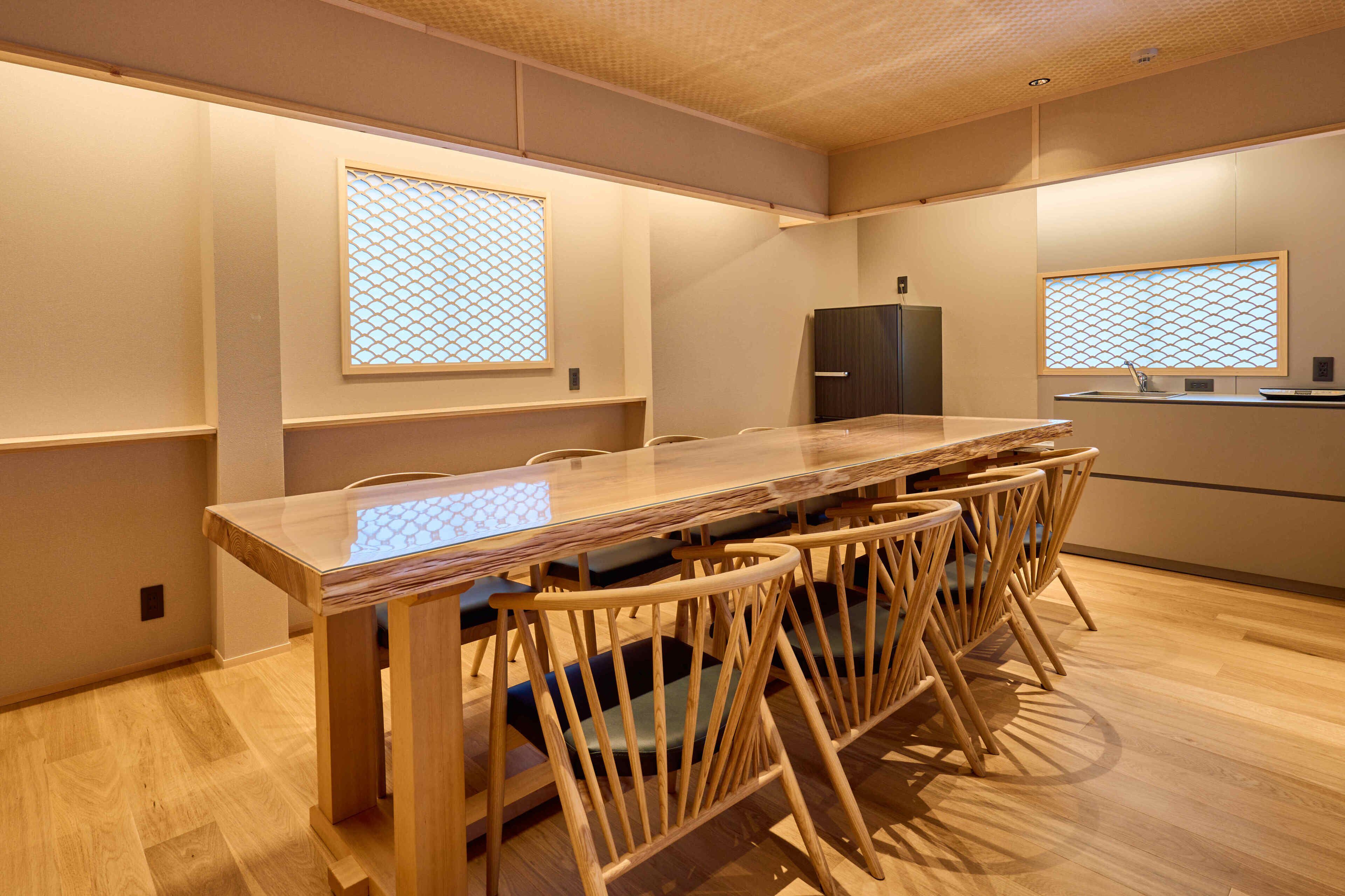 Uma área de refeições moderna com uma mesa de madeira comprida e cadeiras combinando, uma geladeira e duas janelas com painéis decorativos.