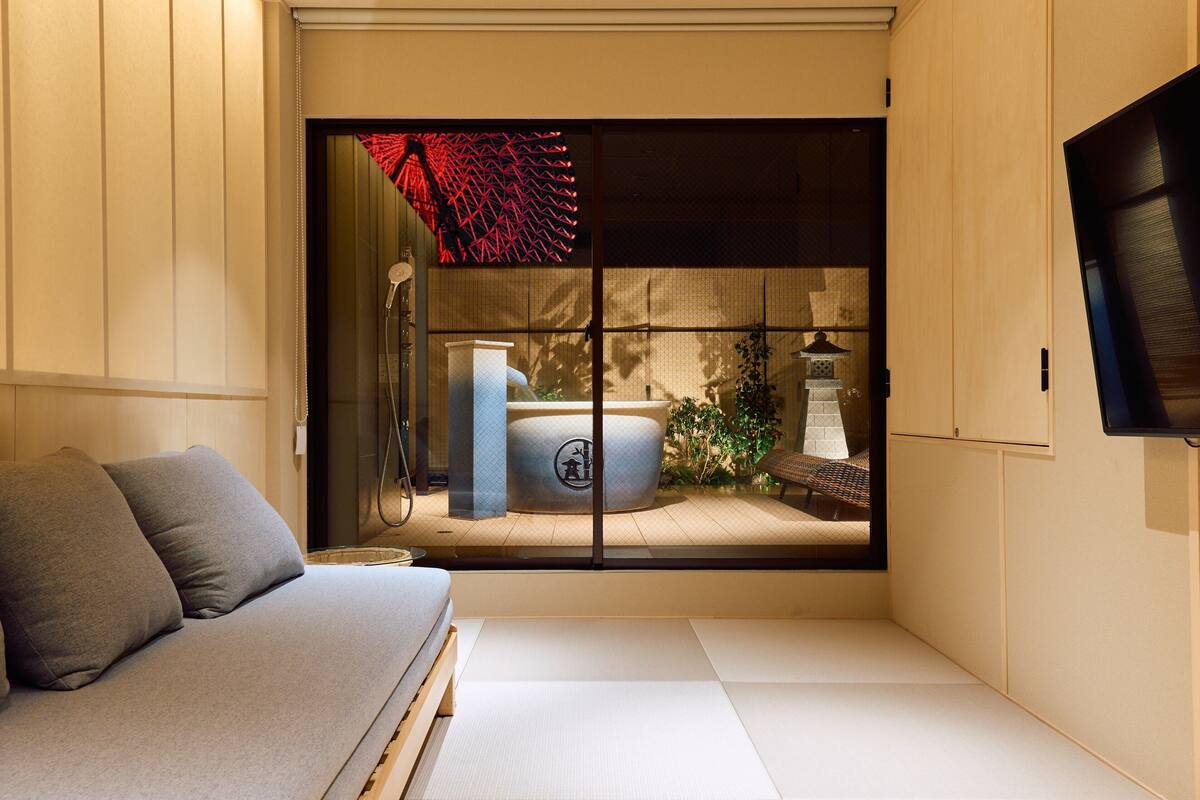 Una habitación minimalista con suelo de tatami, una cama de madera baja, un sofá gris y un panel shoji corredero con un detalle decorativo rojo.