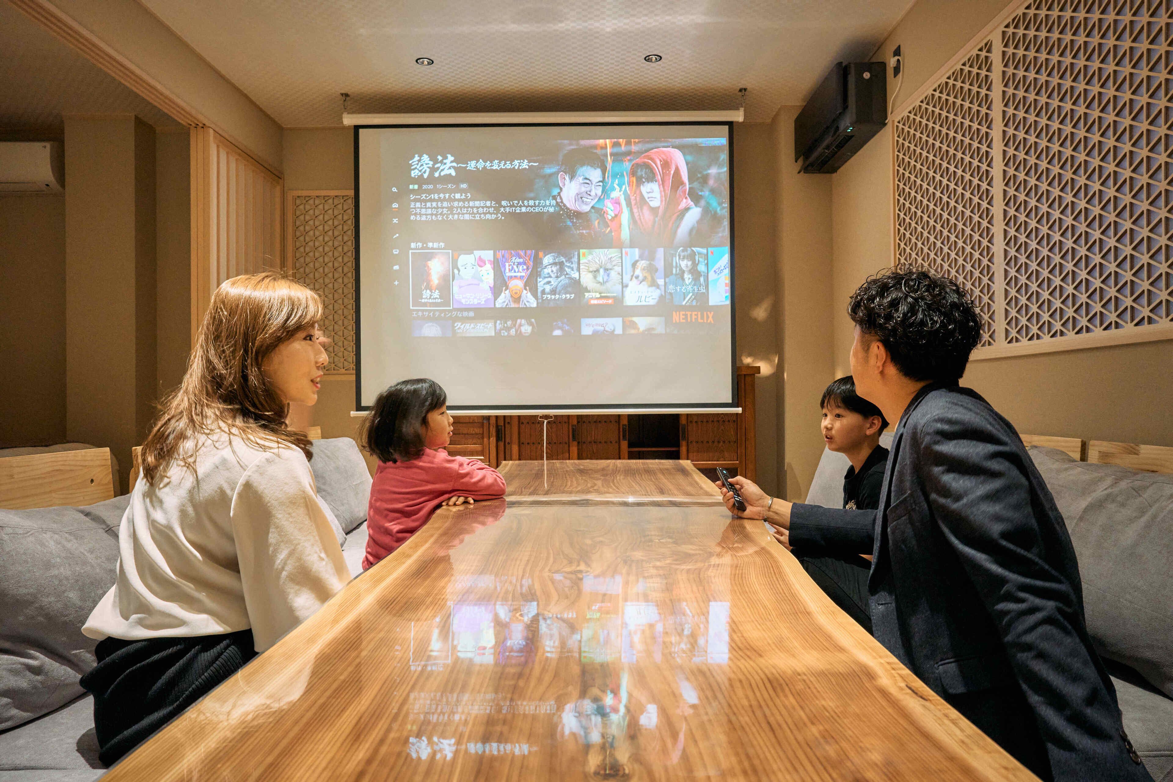 A group of people seated around a wooden table in a room with a projection screen displaying a Netflix interface.