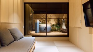 A minimalist room with a tatami mat floor, a low wooden bed, a grey sofa, and a sliding shoji screen door leading to a traditional Japanese bath area with a stone lantern. - KONJAKUSO Osaka Tempozan "IKESU" Projection Mapping SPA "TAKADORO" Wheel View (Osaka)