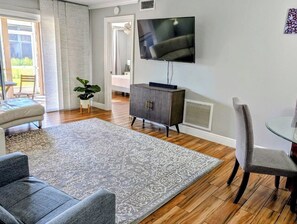 Ein Wohnzimmer mit einem an der Wand befestigten Flachbildfernseher, einem grauen Teppich mit Muster, einer dunklen Holzkommode und einem Essbereich mit einem Glastisch und Stühlen.