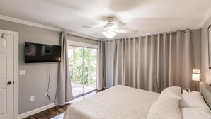 A bedroom with a bed, a ceiling fan, a TV mounted on the wall, and a sliding glass door leading to a balcony with curtains.