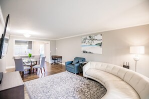 En moderne stue med en buet hvid sofa, en blå lænestol og et spiseområde med et glasbord og stole.