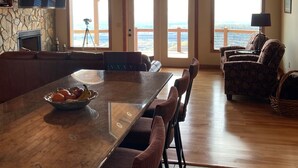 A spacious dining area with a long table and multiple chairs, a stone wall with a flat-screen TV, and large windows offering a view of the outdoors.