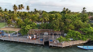 A wooden deck with lounge chairs and a covered seating area overlooking a body of water, surrounded by lush greenery and palm trees.
