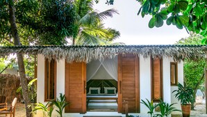 A thatched-roof hut with a bed inside, surrounded by tropical plants and palm trees.