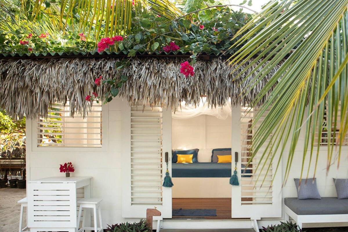 A thatched-roof outdoor seating area with a bed and a table, surrounded by plants and a palm tree.