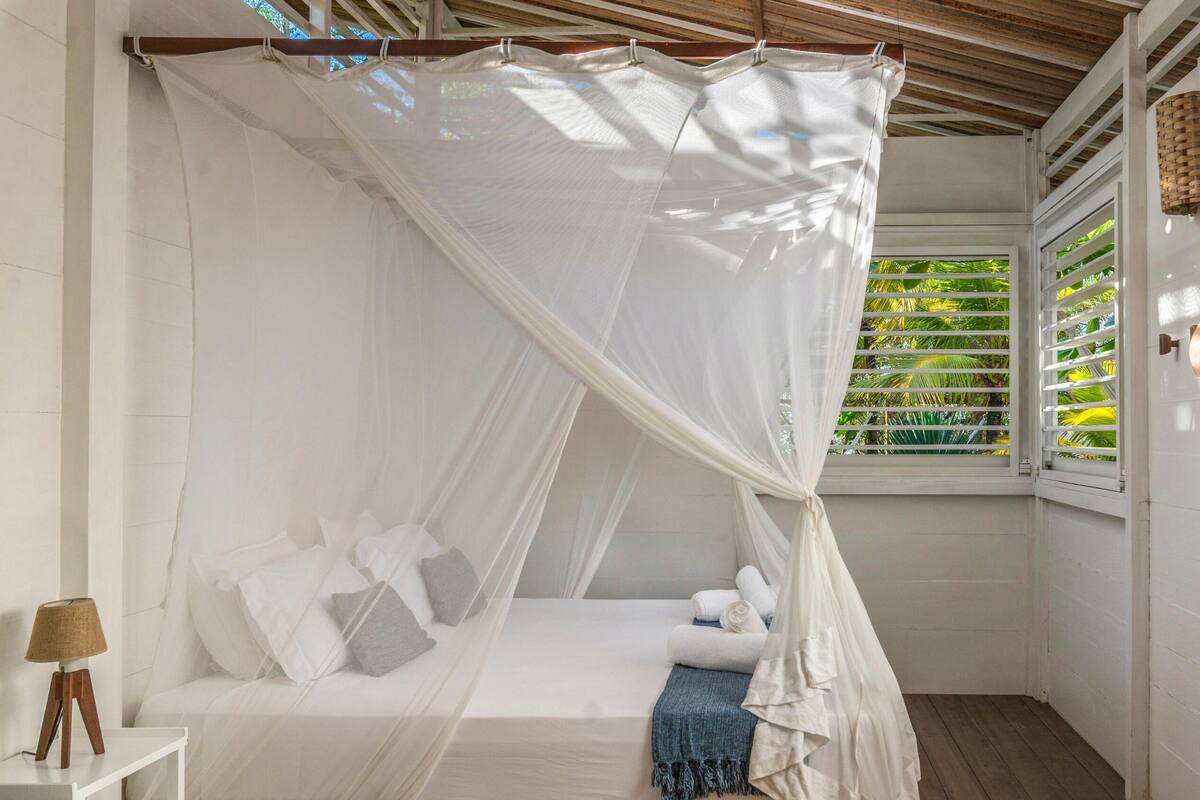 A white four-poster canopy bed with pillows and a rolled towel, a small bedside table with a lamp, and a woven rug on a tiled floor.