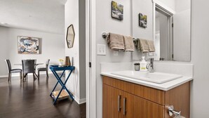 A modern bathroom with a white sink, wooden cabinets, and a large mirror. There are two towels hanging on the wall and a small potted plant on the counter. - TC Condo | Steps to Tart Trail | Near Downtown (Traverse City)