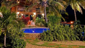 Un edificio de dos pisos con techo de paja, rodeado de palmeras y una piscina.