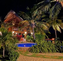 Um edifício de dois andares com telhado de palha, rodeado por palmeiras e uma piscina.