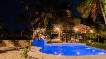 A swimming pool surrounded by palm trees and a stone wall, with a wooden deck and a thatched-roof structure in the background.
