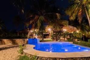 A swimming pool surrounded by palm trees and a stone wall, with a wooden deck and a thatched-roof structure in the background. - Moriá Eco Lodge (Amontada)