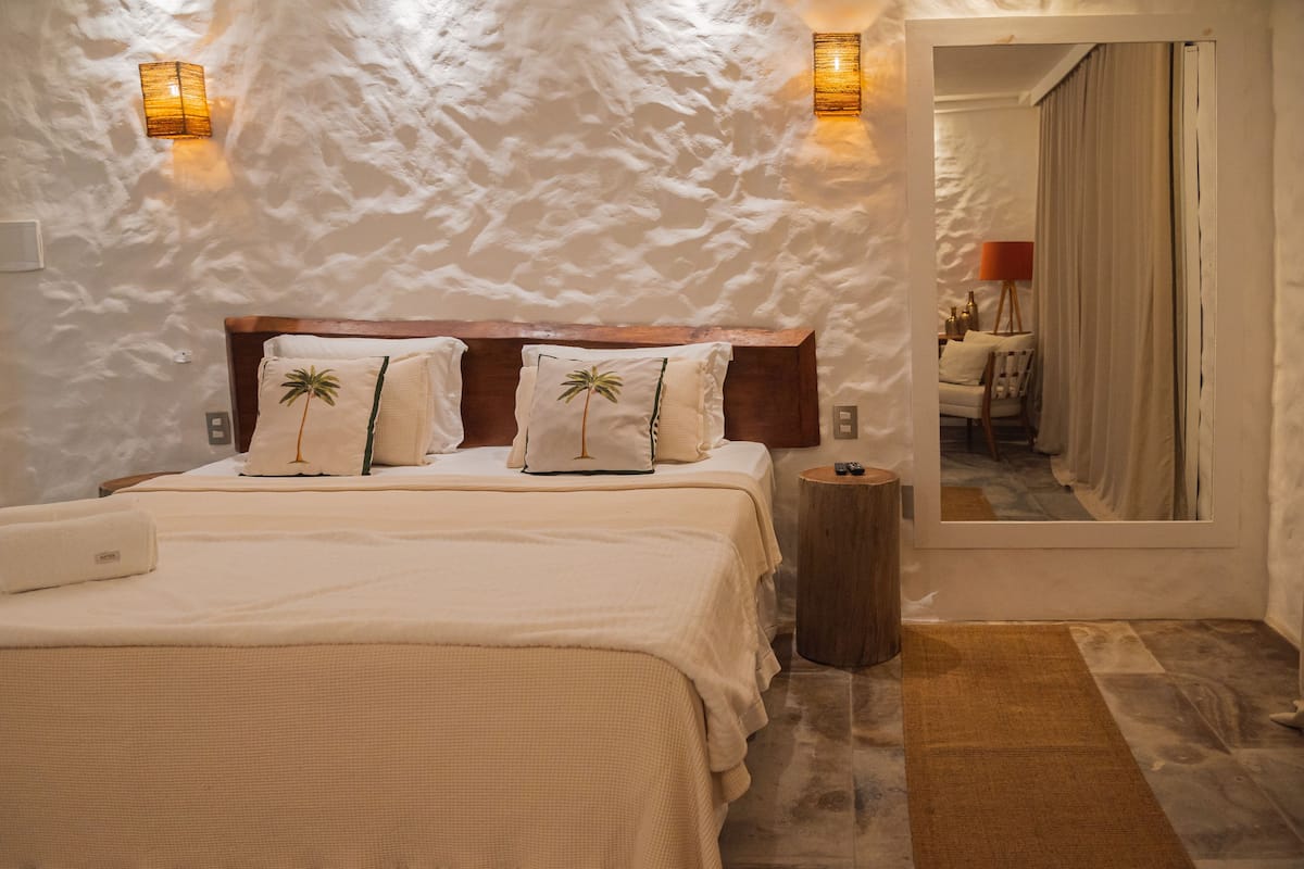 Ein Schlafzimmer mit strukturierter Wand, einem Holzkopfteil, zwei Betten mit passenden Kissen, einem Nachttisch, einem Spiegel und Blick in einen anderen Raum.