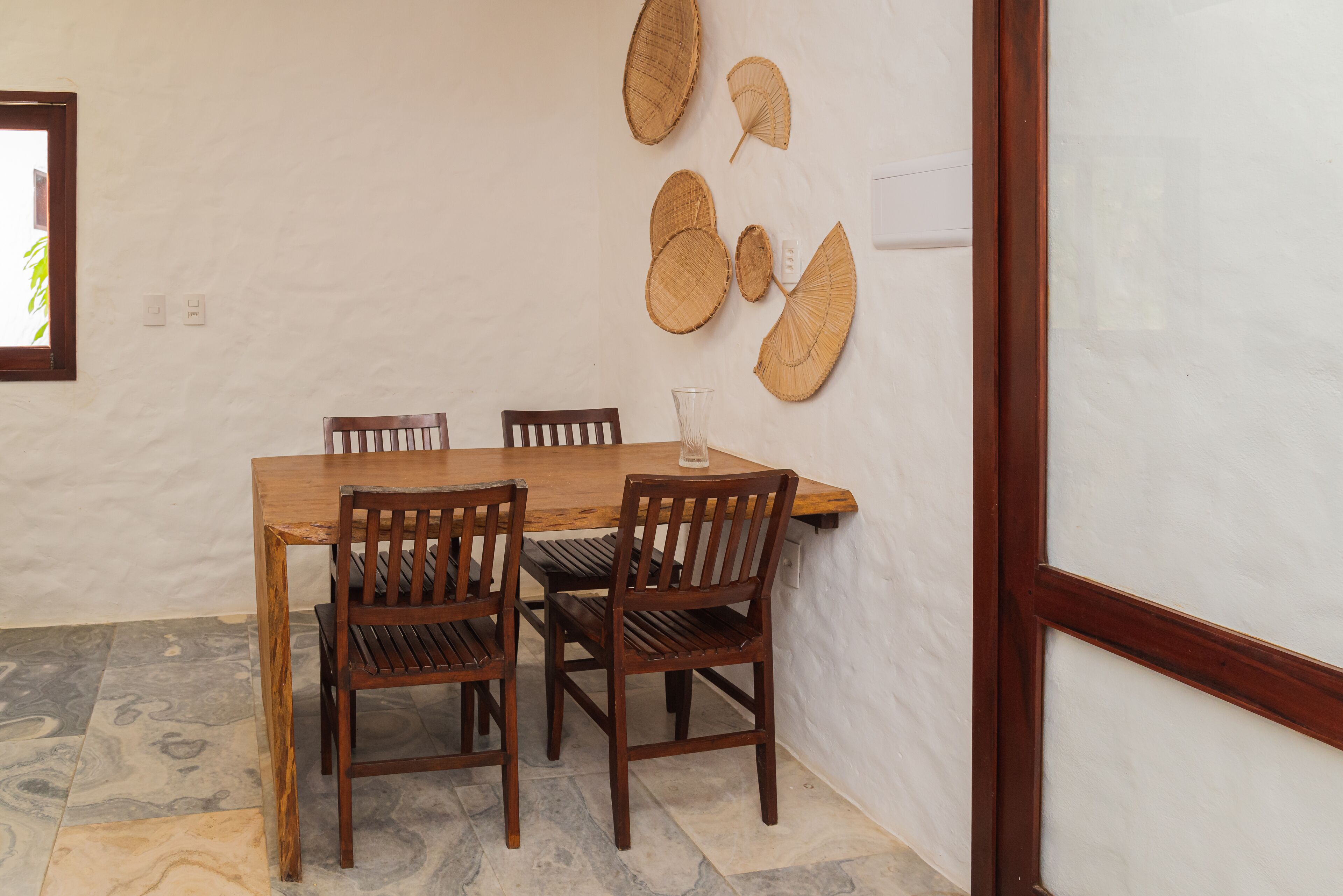 A dining area with a wooden table and four chairs, a glass on the table, and woven wall decorations.