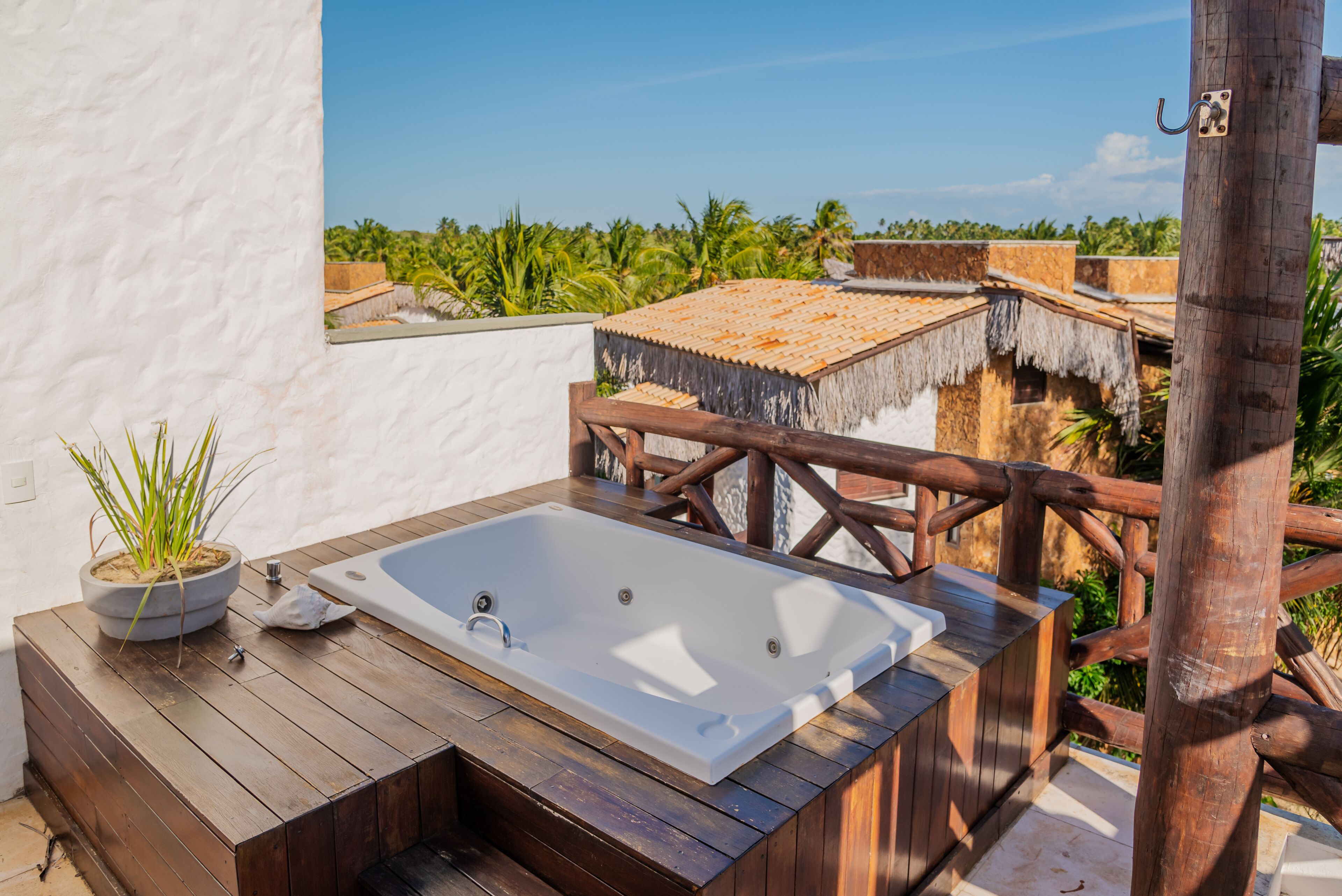 A wooden deck with a hot tub, a potted plant, and a view of a thatched-roof building and palm trees.