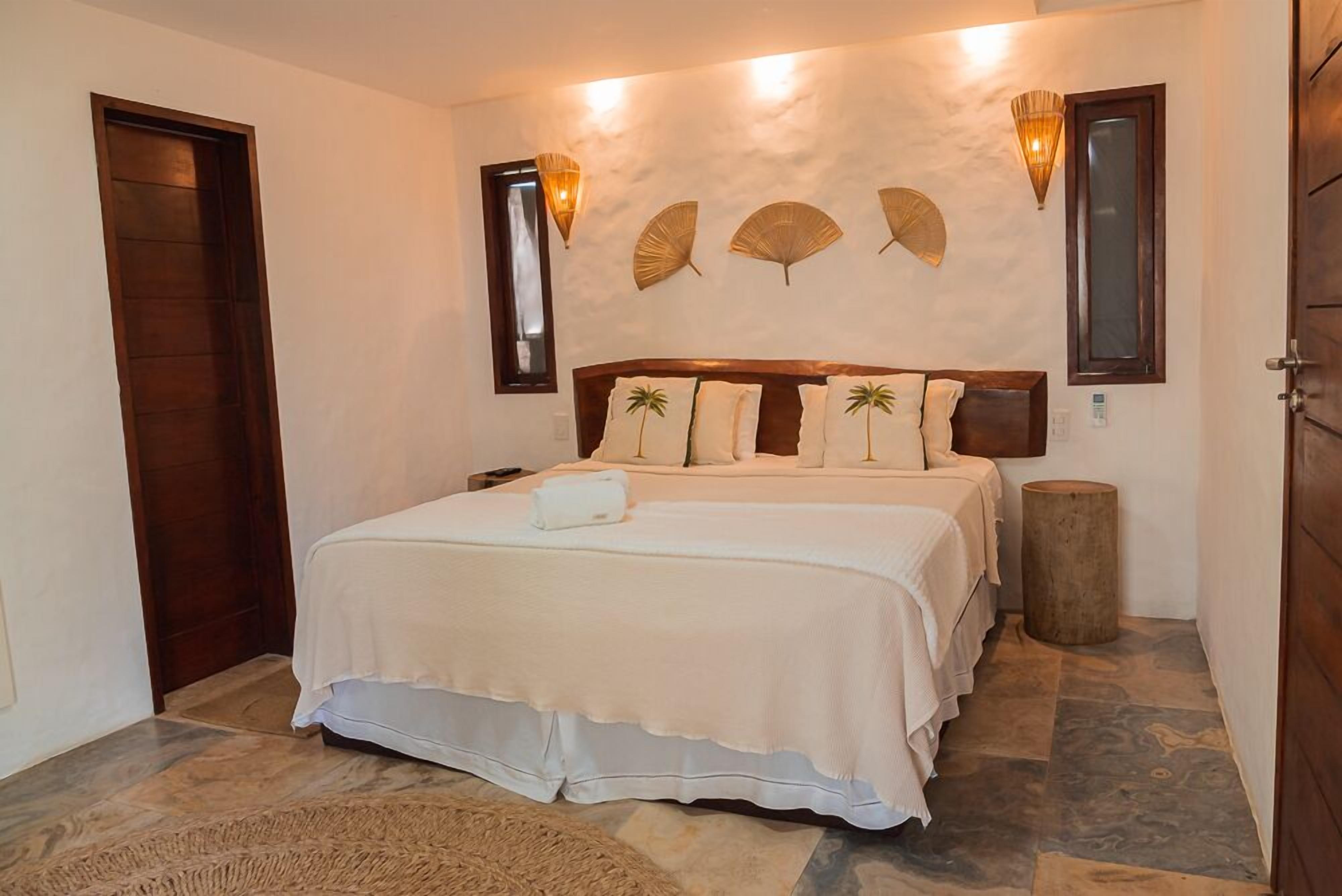 A bedroom with a large bed, two bedside tables, a wooden headboard, wall-mounted lights, and a small round stool.