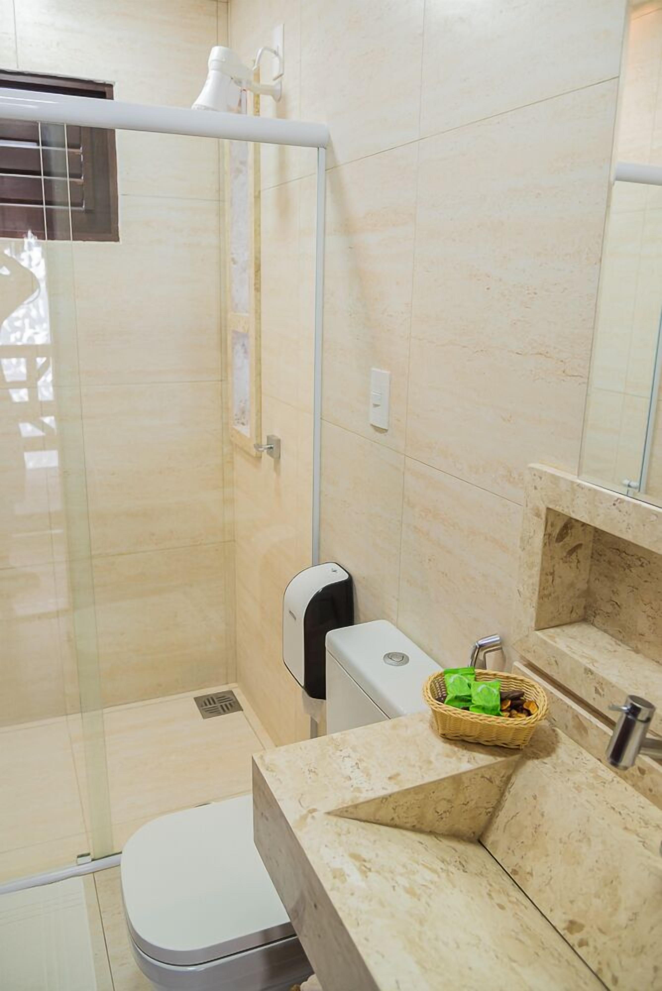 Un baño moderno con cabina de ducha de vidrio, inodoro, lavamanos con encimera de mármol y una cesta pequeña con artículos de aseo.