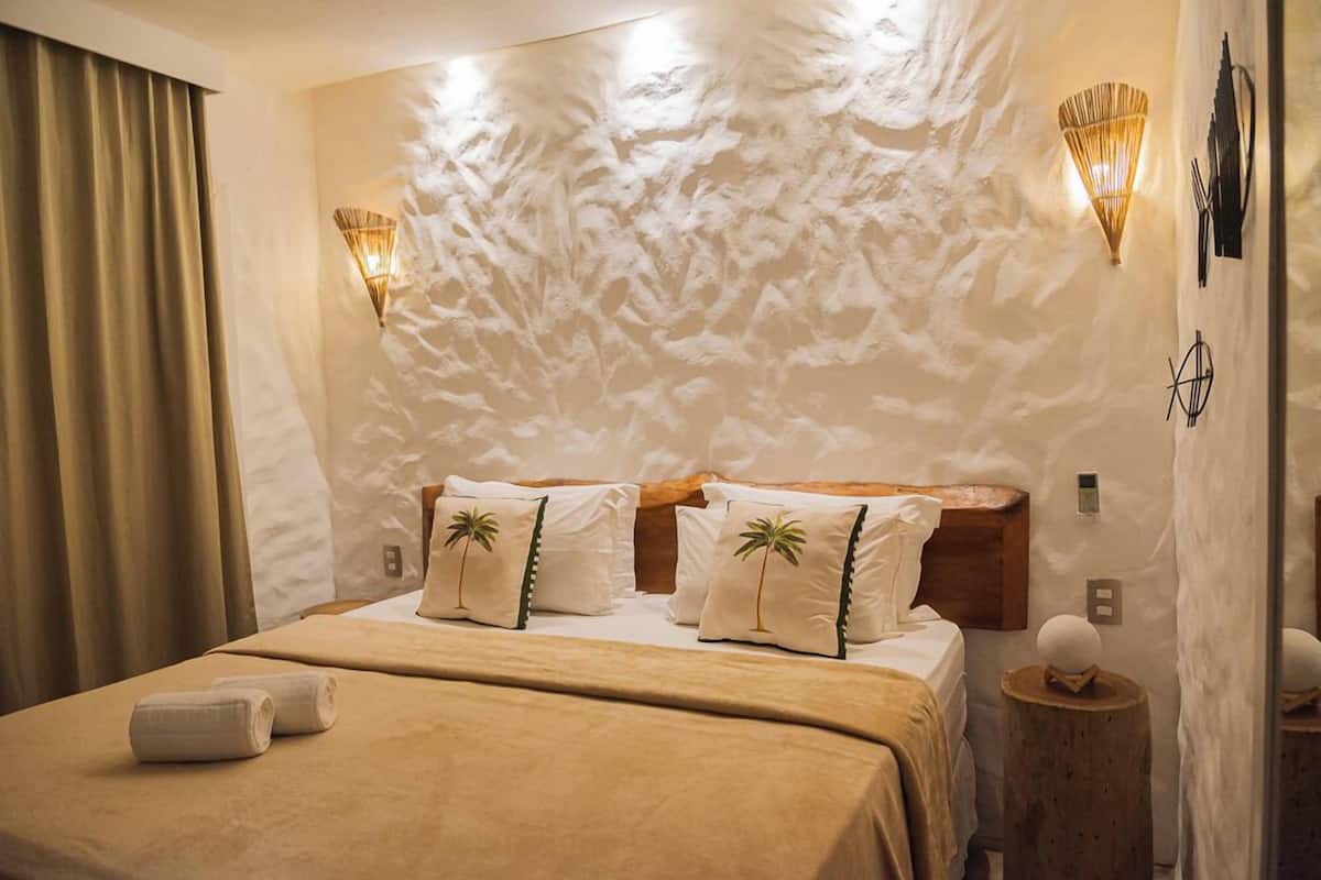 Ein Hotelzimmer mit strukturierter Wand, einem Bett mit beigefarbener Bettwäsche, zwei Palmendruck-Kissen, einem Holz-Kopfteil und zwei Wandleuchten.