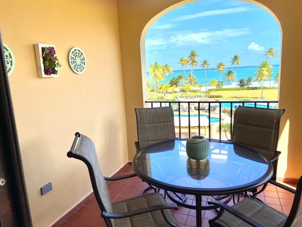 Una mesa redonda de cristal con cuatro sillas, un jarrón decorativo y vistas a una playa con palmeras y una piscina.