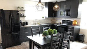 Une cuisine moderne avec des meubles de rangement noirs, un réfrigérateur noir, une table à manger avec des chaises et des luminaires suspendus.