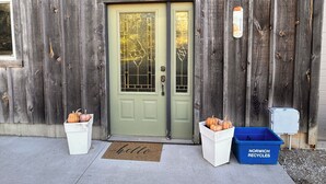 Un bâtiment en bois avec une porte verte, un paillasson où est écrit « Bonjour », deux jardinières blanches contenant des citrouilles et une poubelle de recyclage bleue portant l’inscription « NORWICH RECYCLE ».