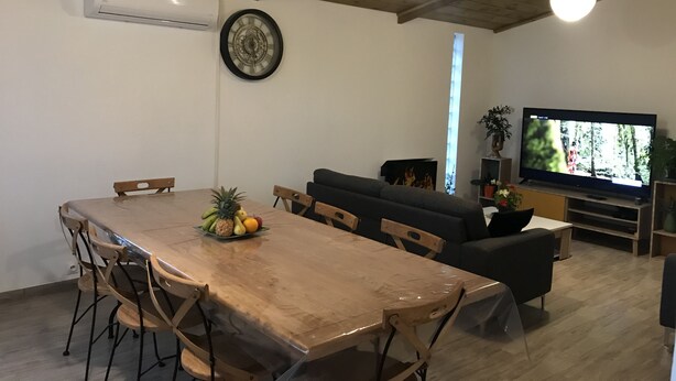 A dining area with a wooden table, chairs, and a fruit centerpiece. There is a sofa, a television, and a fireplace in the background. - Villa de Charme à Bois Court (10 people) (Le Tampon)