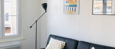 A living room with a black leather sofa, yellow throw blanket, and two polka-dotted pillows. There are two framed pictures on the wall, a floor lamp, and a window with blinds.