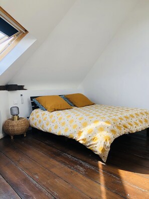 Une chambre avec un plafond en pente, un lit recouvert d’une couvre-lit à motifs floraux jaune et blanc, deux coussins de couleur moutarde, une table d’appoint en osier et une petite lampe.