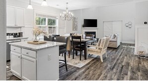 Una cocina moderna con armarios blancos, un comedor con candelabro y una sala de estar con sofá y televisor.