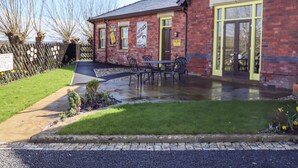 A brick building with a green door and windows, a paved patio with outdoor seating, and a garden with flowers and shrubs.