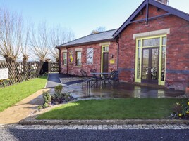 A brick building with a green door and windows, a paved patio with outdoor seating, and a garden with flowers and shrubs.