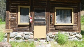 Uma cabana rústica de toras com telhado verde de metal, duas janelas, uma porta e um extintor de incêndio fixado na parede. Uma placa na frente diz 'RHYOLITE'.
