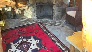 Interior rústico de cabana com lareira de pedra, vigas de madeira, tapete tecido e um ventilador.
