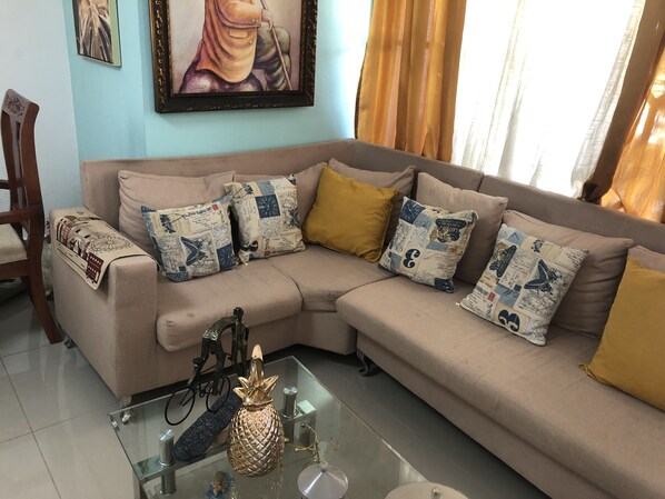 A living room with a beige sectional sofa, patterned throw pillows, a glass coffee table with decorative items, and a framed artwork on the wall.