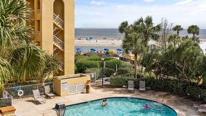 Zona de piscina con vistas a la playa, palmeras y un edificio con balcones.